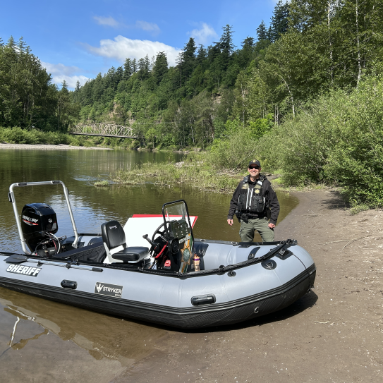 A smiling Multnomah County Sheriff's Office deputy stands on a sandy riverbank next to a grey inflatable patrol boat marked "SHERIFF." The calm river is flanked by a dense forest, with a historic steel truss bridge visible in the background under a blue sky.