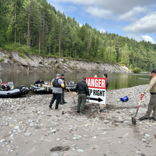 A wide-angle shot shows law enforcement personnel and workers installing a large white and red sign that reads "DANGER KEEP RIGHT" on a rocky river island. Three motorized patrol boats are moored on the riverbank to the left against a backdrop of steep, tree-covered hills.
