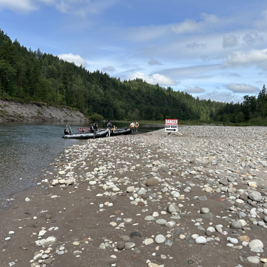 A long shot captures an expansive gravel river bar where a prominent warning sign reading "DANGER KEEP RIGHT" has been installed near the water's edge. Two grey patrol boats are moored nearby on the shore, surrounded by a dense, rolling green forest under a cloudy sky.