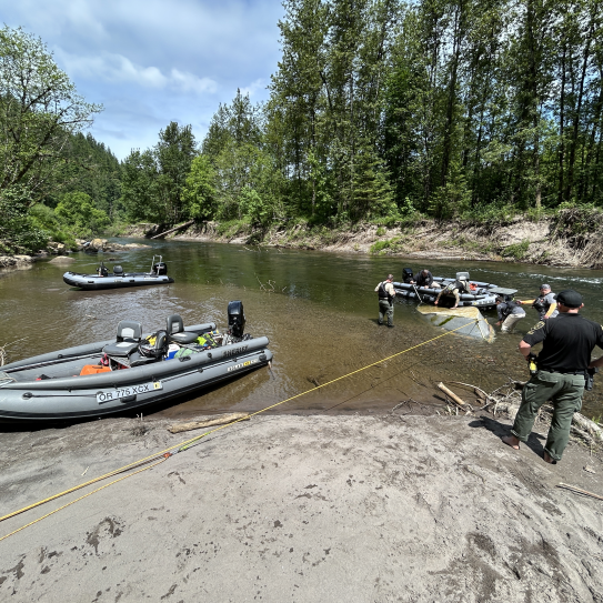 A wide shot from a sandy bank shows several law enforcement members working from multiple grey patrol boats to salvage a submerged drift boat in a calm river. A MCSO deputy stands on the foreground shore monitoring the operation via a yellow tow line attached to the vessel.