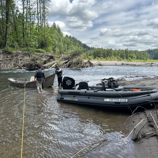 Two crew members stand waist-deep in a river channel to secure and guide a recovered drift boat next to a stationary grey inflatable patrol boat. The background shows a large logjam and river debris along a gravel bar under a cloudy sky.