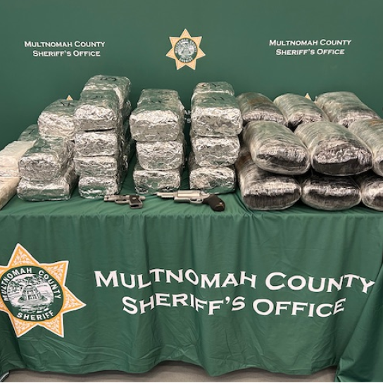 Large amount of illegal narcotics wrapped in plastic on a table with two firearms. Green background with Multnomah County Sheriff's Office logo.