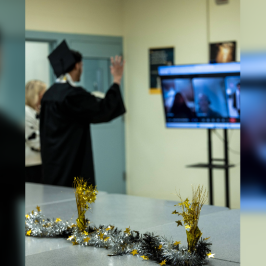 An individual in a graduation gown and cap waves toward a video screen during a virtual ceremony, with festive gold and silver decorations on the table in the foreground.