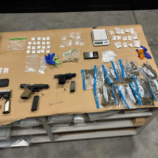 Evidence gathered during a law enforcement investigation is displayed on a large table covered in brown paper. The items include three handguns—one silver and black, one all black with an extended magazine, and one purple and black—alongside loose ammunition and a smartphone. The display also contains a large quantity of narcotics in various types of packaging: a cluster of blue-sealed plastic bags with green plant material, dozens of small clear baggies with white powder or crystals, and a larger bag.
