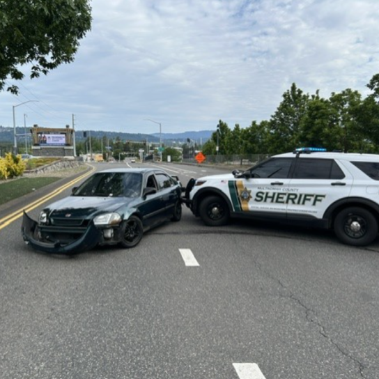 Side profile scene showing a white and green Multnomah County Sheriff’s Office patrol SUV utilizing its front push bumper to pin the rear driver-side quarter panel of a dark green Honda sedan. The Honda is positioned sideways across the roadway lanes with its front bumper completely detached and resting on the asphalt. The patrol vehicle’s emergency lights are active, and the background shows a curved road leading toward a highway interchange with trees and distant hills under an overcast sky.