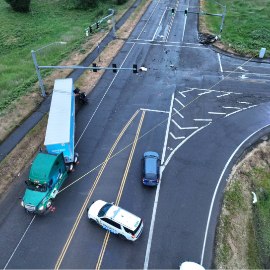 A drone photograph captures the scene of a fatal two-vehicle collision at a wide, multi-lane T-intersection. A green commercial semi-truck with a blue trailer is stopped along the left shoulder near a patrol car, while debris is scattered across the dark, wet asphalt leading toward a severely burned vehicle on the far right.