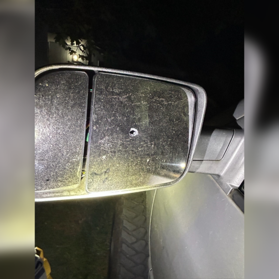 A close-up, nighttime photograph of a dusty, silver truck's passenger-side towing mirror showing a single, clean bullet hole punctured directly through the center of the glass pane.