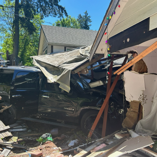 A black SUV crashed through the side of a house, causing extensive structural damage to the wall and roof. Debris, including broken wood and insulation, is scattered around the vehicle. Multicolored holiday lights are still strung along the damaged roofline.