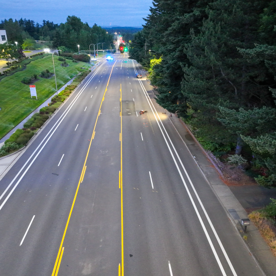 A high-angle drone photograph showing an empty, multi-lane road bordered by manicured green lawns and parking lots on the left and a thick canopy of pine trees on the right. A single motorcycle is down in the right-hand lane, and an MCSO patrol SUV blocks the road further in the distance.