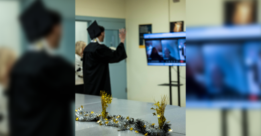 An individual in a graduation gown and cap waves toward a video screen during a virtual ceremony, with festive gold and silver decorations on the table in the foreground.