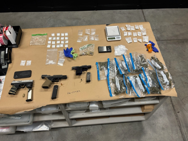 Evidence gathered during a law enforcement investigation is displayed on a large table covered in brown paper. The items include three handguns—one silver and black, one all black with an extended magazine, and one purple and black—alongside loose ammunition and a smartphone. The display also contains a large quantity of narcotics in various types of packaging: a cluster of blue-sealed plastic bags with green plant material, dozens of small clear baggies with white powder or crystals, and a larger bag.