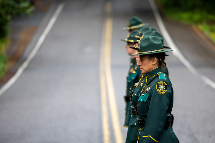 A line of Multnomah County Sheriff's Office deputies assigned to the MCSO Honor Guard stand in a disciplined row on a paved road. They are dressed in formal green uniforms with gold braiding, hats, and agency patches. The deputies are shown in profile, looking forward with solemn expressions, while the road behind them stretches into a soft, out-of-focus background.