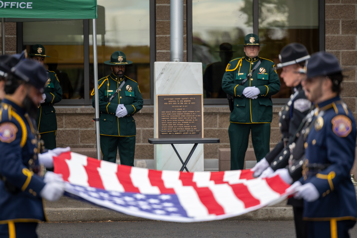 MCSO Honor Guard members in green dress uniforms stand at attention behind a memorial plaque. In the foreground, personnel from partner agencies in navy uniforms hold an unfurled American flag during the solemn ceremony.