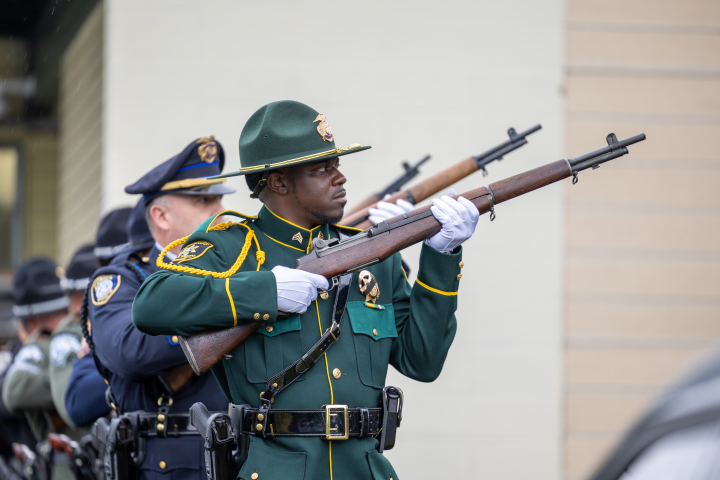 A close-up profile of an MCSO Honor Guard member in a forest green dress uniform and hat, holding a rifle at an angle during a 21-gun salute. He wears white gloves and gold braiding, with other uniformed personnel lined up behind him in a soft-focus background.