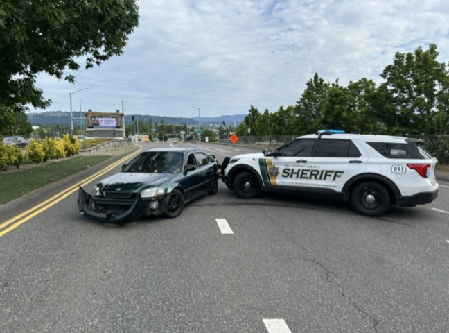 Side profile scene showing a white and green Multnomah County Sheriff’s Office patrol SUV utilizing its front push bumper to pin the rear driver-side quarter panel of a dark green Honda sedan. The Honda is positioned sideways across the roadway lanes with its front bumper completely detached and resting on the asphalt. The patrol vehicle’s emergency lights are active, and the background shows a curved road leading toward a highway interchange with trees and distant hills under an overcast sky.