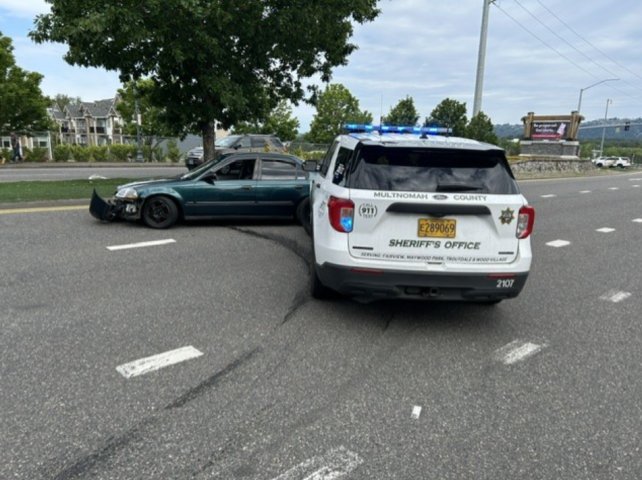 Image: A white Multnomah County Sheriff’s Office Ford Explorer patrol SUV with flashing blue emergency lights is stopped at an angle behind a dark green Honda sedan on a multi-lane asphalt road. Heavy black tire skid marks curve across the asphalt lanes, tracing the path where the Honda was spun sideways following a Pursuit Intervention Technique (PIT) maneuver. In the background, a landscaped median, trees, and a roadside digital billboard are visible under a cloudy sky.