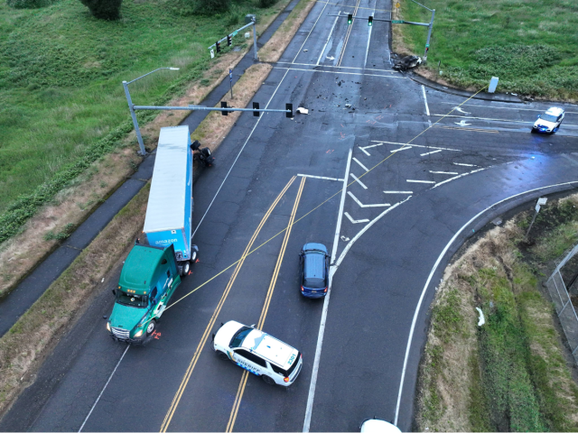 A drone photograph captures the scene of a fatal two-vehicle collision at a wide, multi-lane T-intersection. A green commercial semi-truck with a blue trailer is stopped along the left shoulder near a patrol car, while debris is scattered across the dark, wet asphalt leading toward a severely burned vehicle on the far right.