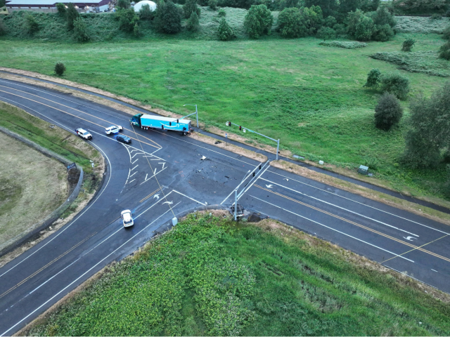 An aerial view shows a broad look at an intersection cordoned off by yellow crime scene tape and multiple patrol SUVs. A commercial semi-truck sits angled across the upper lanes, and a charred vehicle rests off the shoulder on the right, surrounded by green fields.