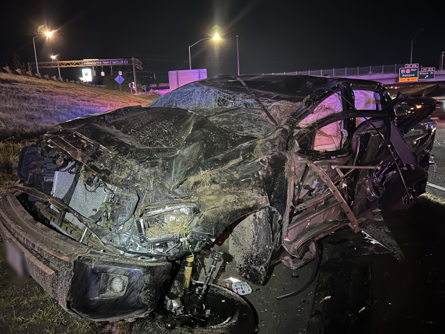A close up from the hood of a black pickup truck that rolled over with the front windshield damaged and heavy front-end damage after rolling on the freeway. 
