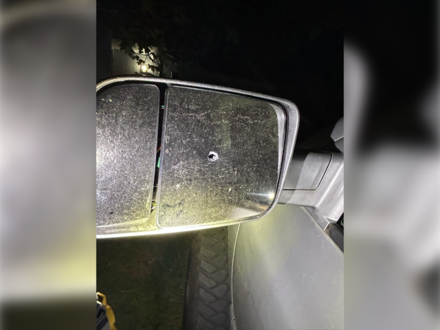 A close-up, nighttime photograph of a dusty, silver truck's passenger-side towing mirror showing a single, clean bullet hole punctured directly through the center of the glass pane.