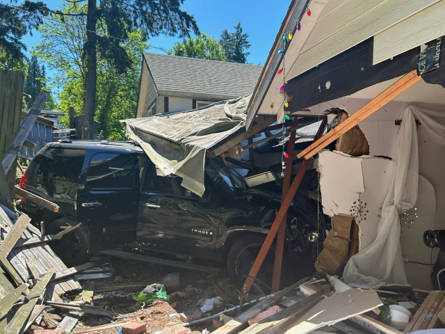 A black SUV crashed through the side of a house, causing extensive structural damage to the wall and roof. Debris, including broken wood and insulation, is scattered around the vehicle. Multicolored holiday lights are still strung along the damaged roofline.