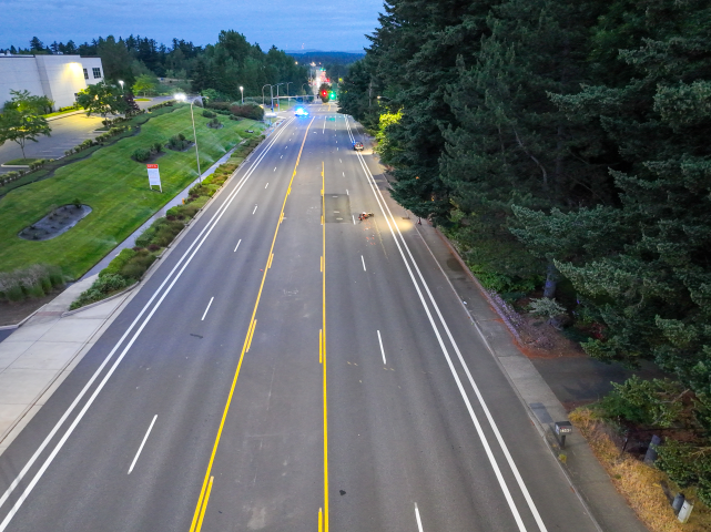 A high-angle drone photograph showing an empty, multi-lane road bordered by manicured green lawns and parking lots on the left and a thick canopy of pine trees on the right. A single motorcycle is down in the right-hand lane, and an MCSO patrol SUV blocks the road further in the distance.