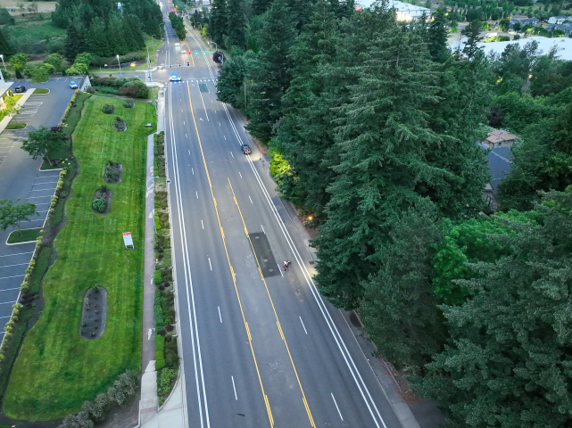 A drone view looking down a multi-lane roadway at dawn. A motorcycle lies on its side in the far right lane next to a dense line of tall evergreen trees, while an MCSO vehicle with flashing blue lights blocks the distant intersection.