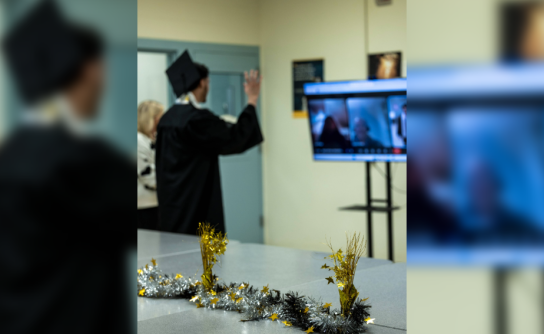 An individual in a graduation gown and cap waves toward a video screen during a virtual ceremony, with festive gold and silver decorations on the table in the foreground.