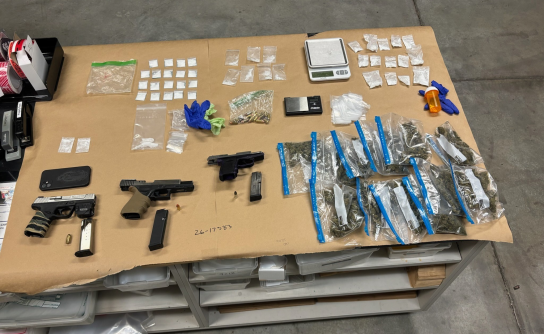Evidence gathered during a law enforcement investigation is displayed on a large table covered in brown paper. The items include three handguns—one silver and black, one all black with an extended magazine, and one purple and black—alongside loose ammunition and a smartphone. The display also contains a large quantity of narcotics in various types of packaging: a cluster of blue-sealed plastic bags with green plant material, dozens of small clear baggies with white powder or crystals, and a larger bag.
