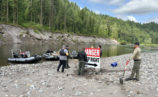 A wide-angle shot shows law enforcement personnel and workers installing a large white and red sign that reads "DANGER KEEP RIGHT" on a rocky river island. Three motorized patrol boats are moored on the riverbank to the left against a backdrop of steep, tree-covered hills.