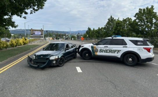 Side profile scene showing a white and green Multnomah County Sheriff’s Office patrol SUV utilizing its front push bumper to pin the rear driver-side quarter panel of a dark green Honda sedan. The Honda is positioned sideways across the roadway lanes with its front bumper completely detached and resting on the asphalt. The patrol vehicle’s emergency lights are active, and the background shows a curved road leading toward a highway interchange with trees and distant hills under an overcast sky.