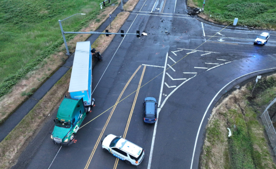 A drone photograph captures the scene of a fatal two-vehicle collision at a wide, multi-lane T-intersection. A green commercial semi-truck with a blue trailer is stopped along the left shoulder near a patrol car, while debris is scattered across the dark, wet asphalt leading toward a severely burned vehicle on the far right.