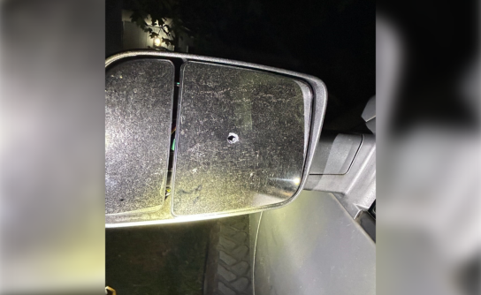 A close-up, nighttime photograph of a dusty, silver truck's passenger-side towing mirror showing a single, clean bullet hole punctured directly through the center of the glass pane.