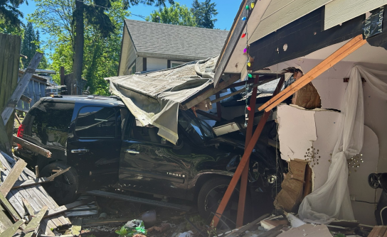 A black SUV crashed through the side of a house, causing extensive structural damage to the wall and roof. Debris, including broken wood and insulation, is scattered around the vehicle. Multicolored holiday lights are still strung along the damaged roofline.