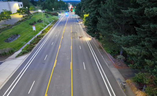 A high-angle drone photograph showing an empty, multi-lane road bordered by manicured green lawns and parking lots on the left and a thick canopy of pine trees on the right. A single motorcycle is down in the right-hand lane, and an MCSO patrol SUV blocks the road further in the distance.